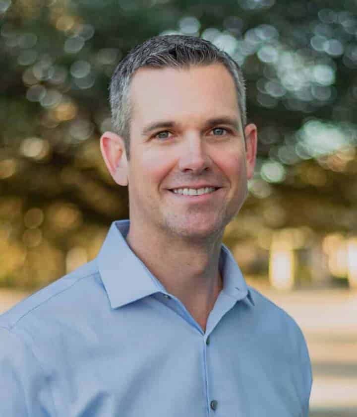 Man with short dark hair and a light blue collared shirt smiles while standing outdoors, with trees and sunlight blurred in the background.
