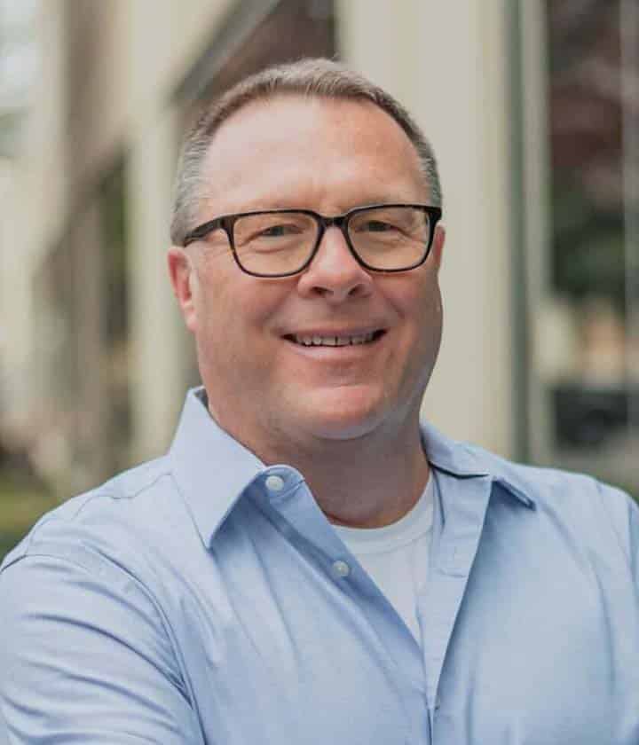 A man with short hair and glasses, wearing a light blue button-up shirt over a white t-shirt, stands outdoors in front of a building, smiling at the camera.