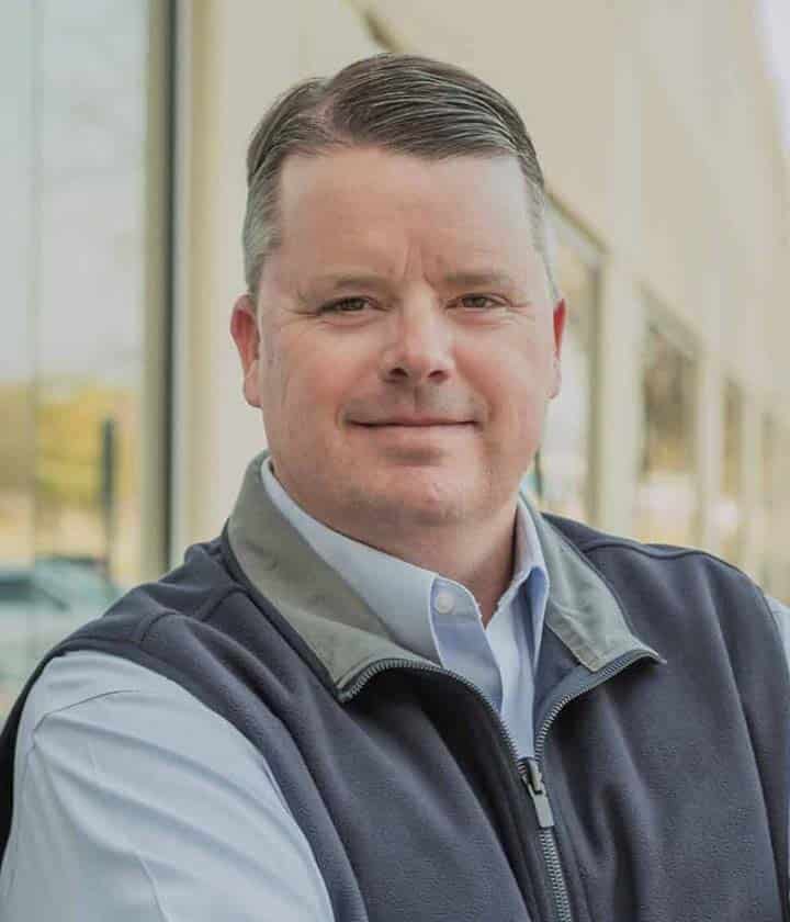 A man with short brown hair wearing a light blue shirt and a dark vest stands outdoors in front of a building with glass windows.