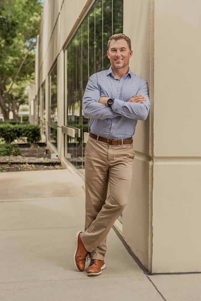 A man in a light blue shirt and khaki pants stands with arms crossed, leaning against a building corner on a sidewalk.