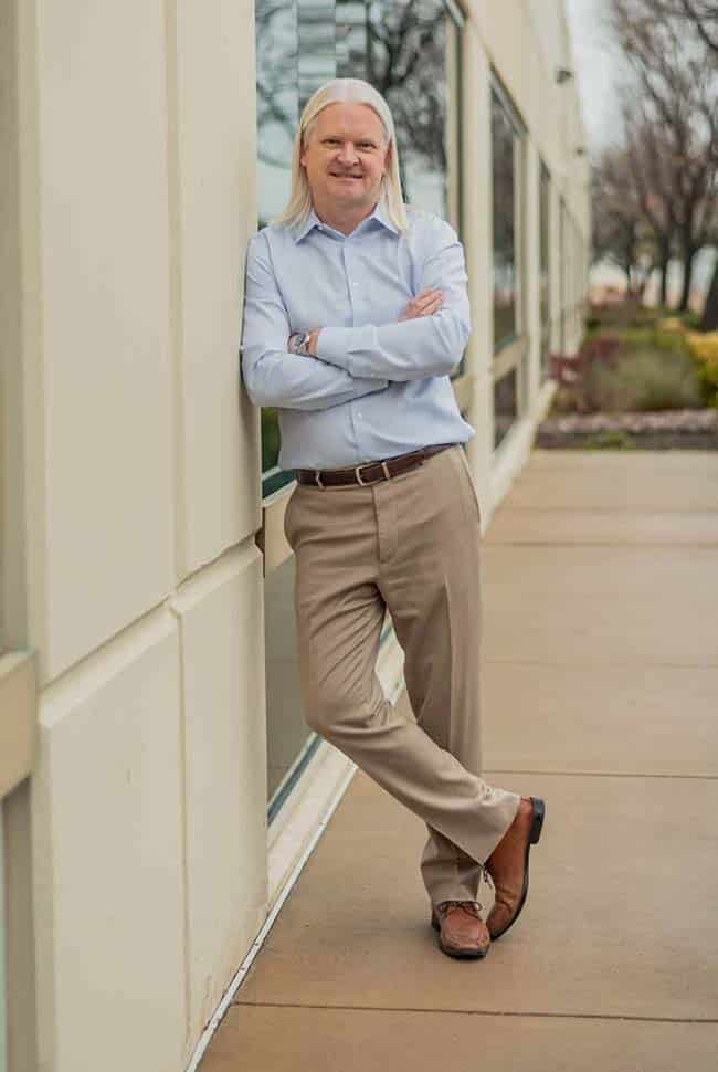 A man with long white hair, wearing a light blue shirt and beige pants, stands with arms crossed, leaning against a building outdoors.