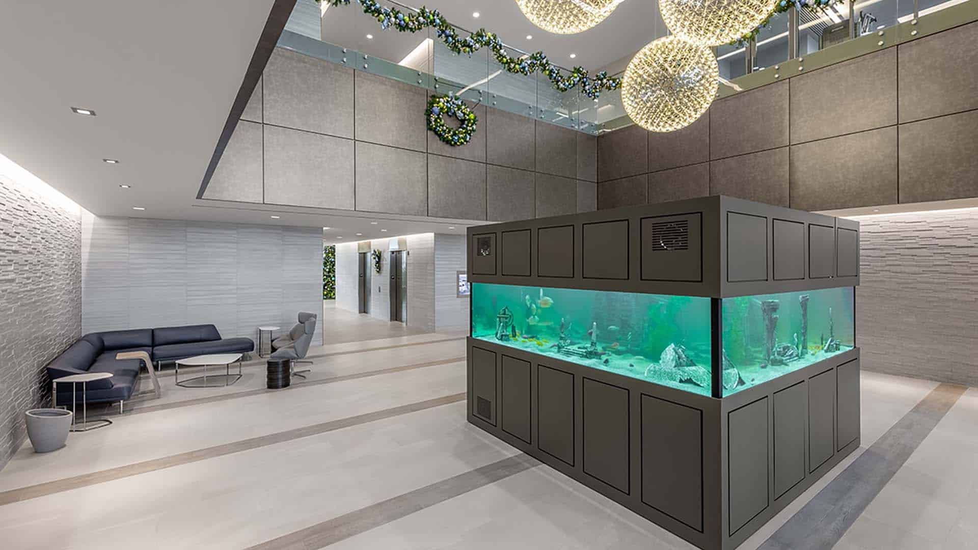 Modern lobby with a large rectangular aquarium in the center, gray seating area on the left, and decorative lights and greenery hanging from the ceiling.