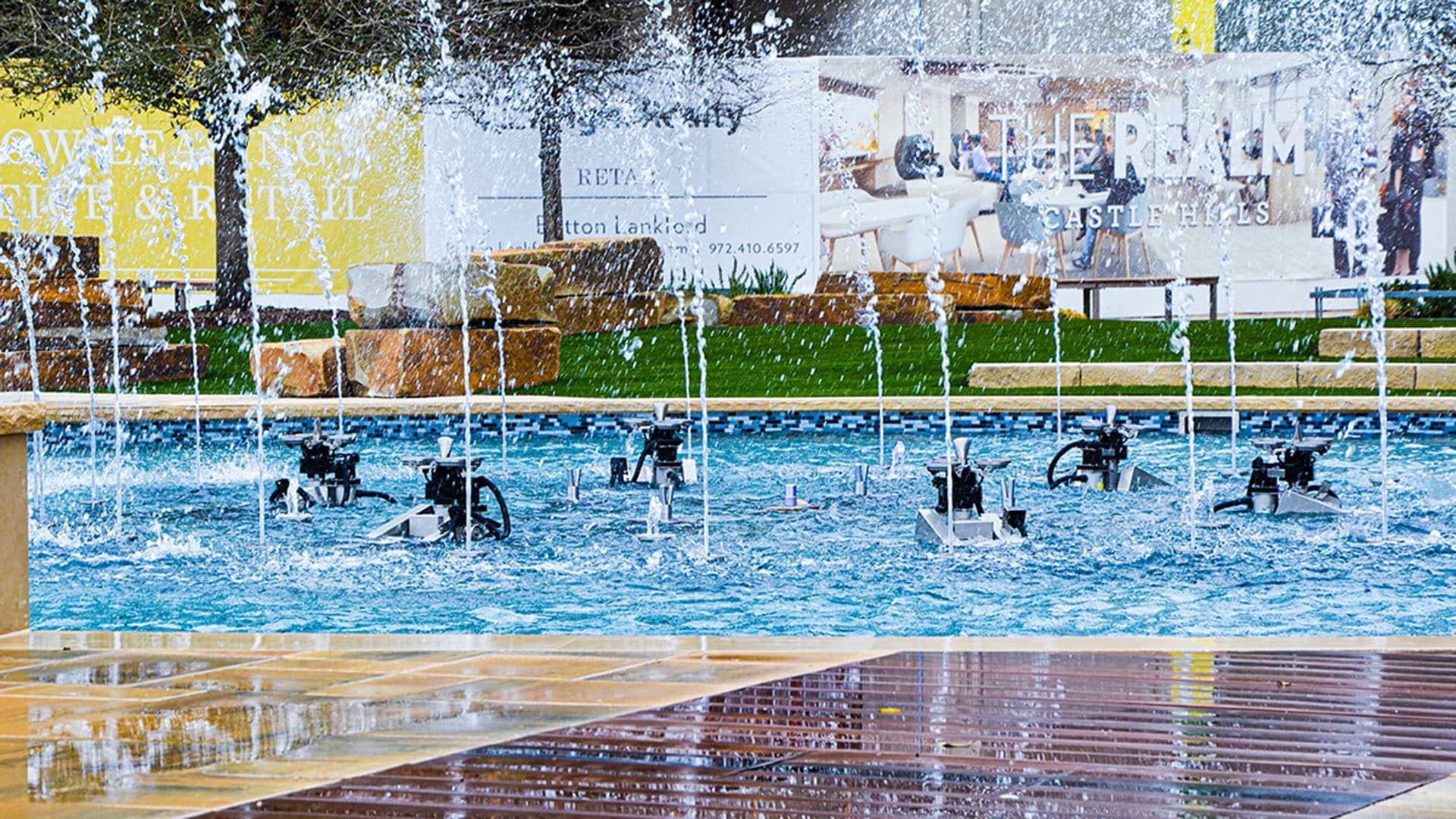 A close-up view of a water fountain with multiple jets spraying water upward, surrounded by wet wooden decking and commercial signs in the background.