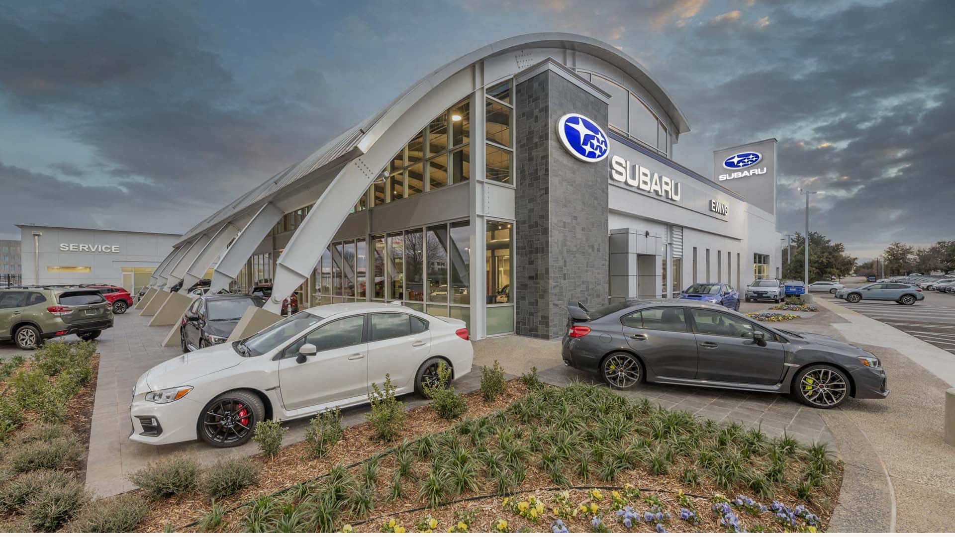 Two Subaru cars are parked in front of a modern Subaru dealership building with glass walls and prominent Subaru signage.