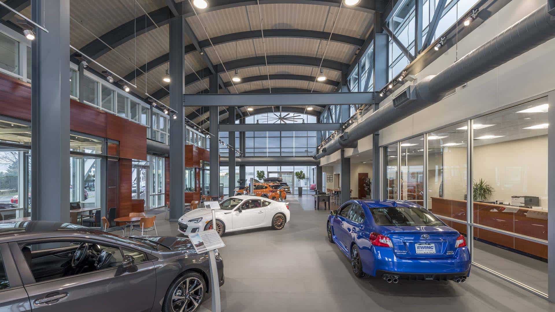 Interior of a modern car dealership showroom with several cars on display, large windows, high ceilings, and office spaces in the background.