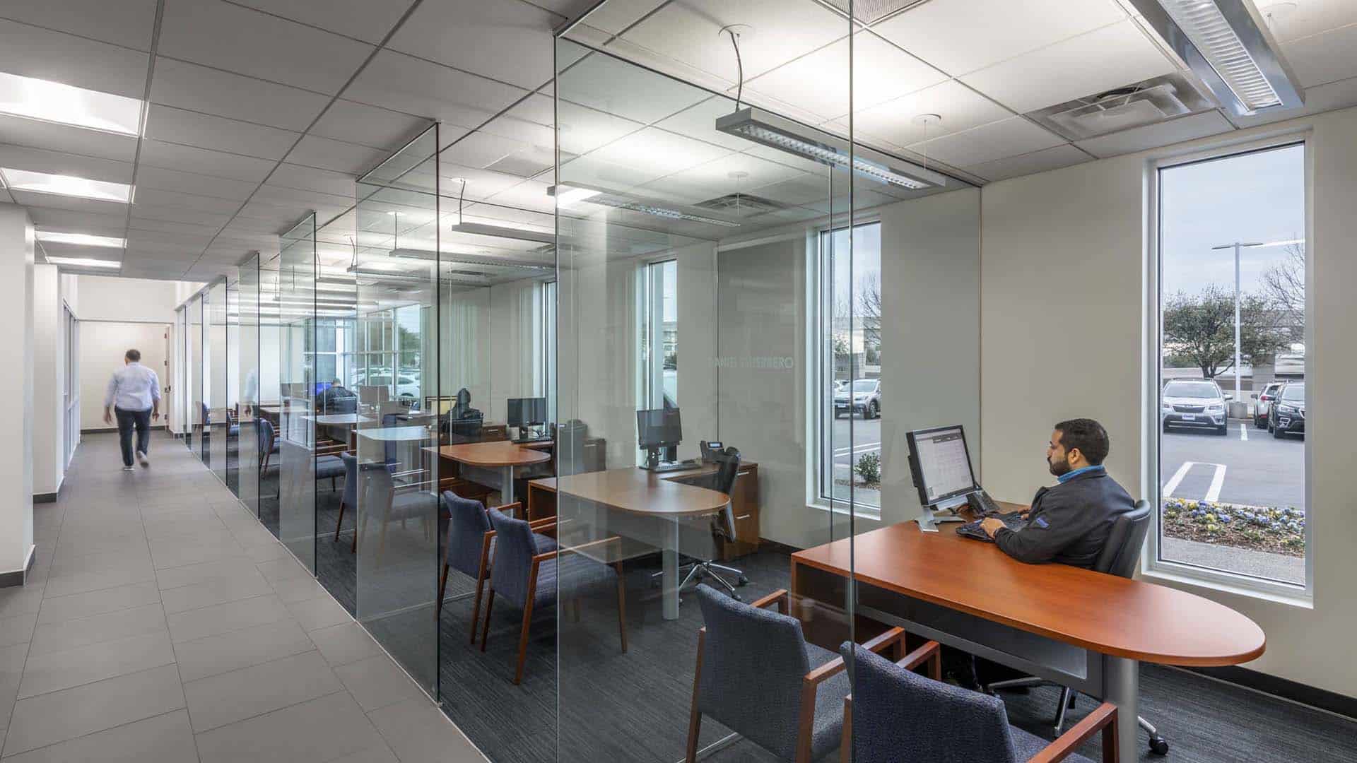 A man works at a computer in a glass-walled office with several desks; another person walks down the hallway. Large windows show a parking lot and trees outside.