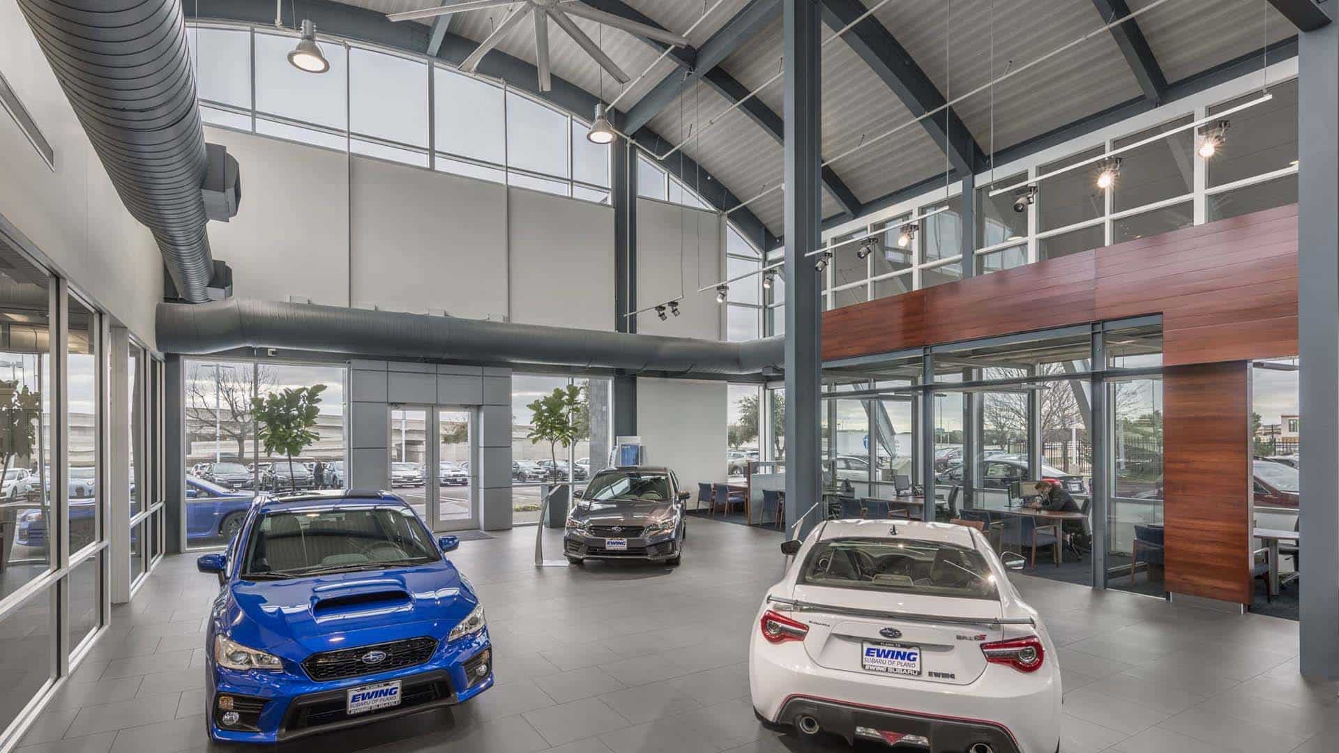 Modern car dealership showroom with large windows, two parked cars in the foreground, a sales office area to the right, and visible cars outside through the glass.