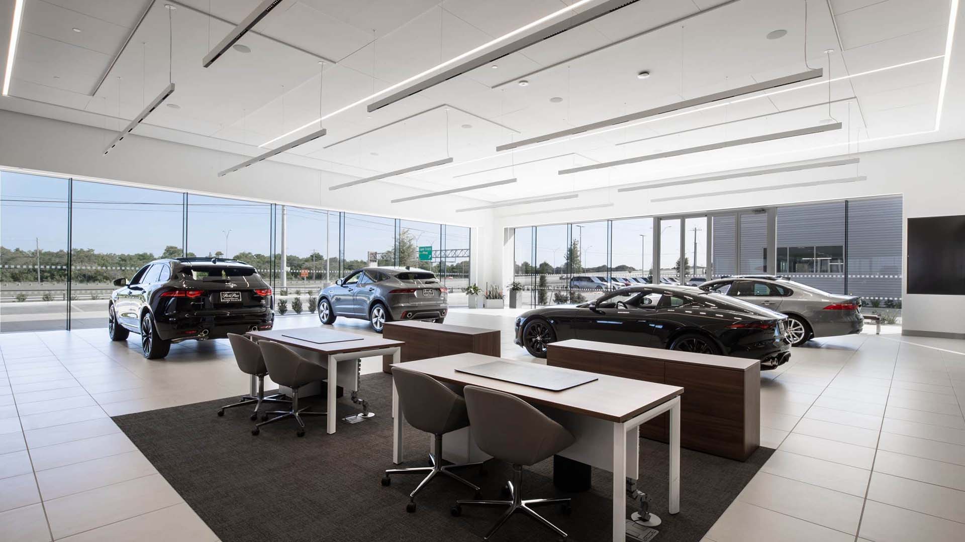 Car showroom interior with four cars on display, two desks with chairs in the foreground, and large windows providing natural light.