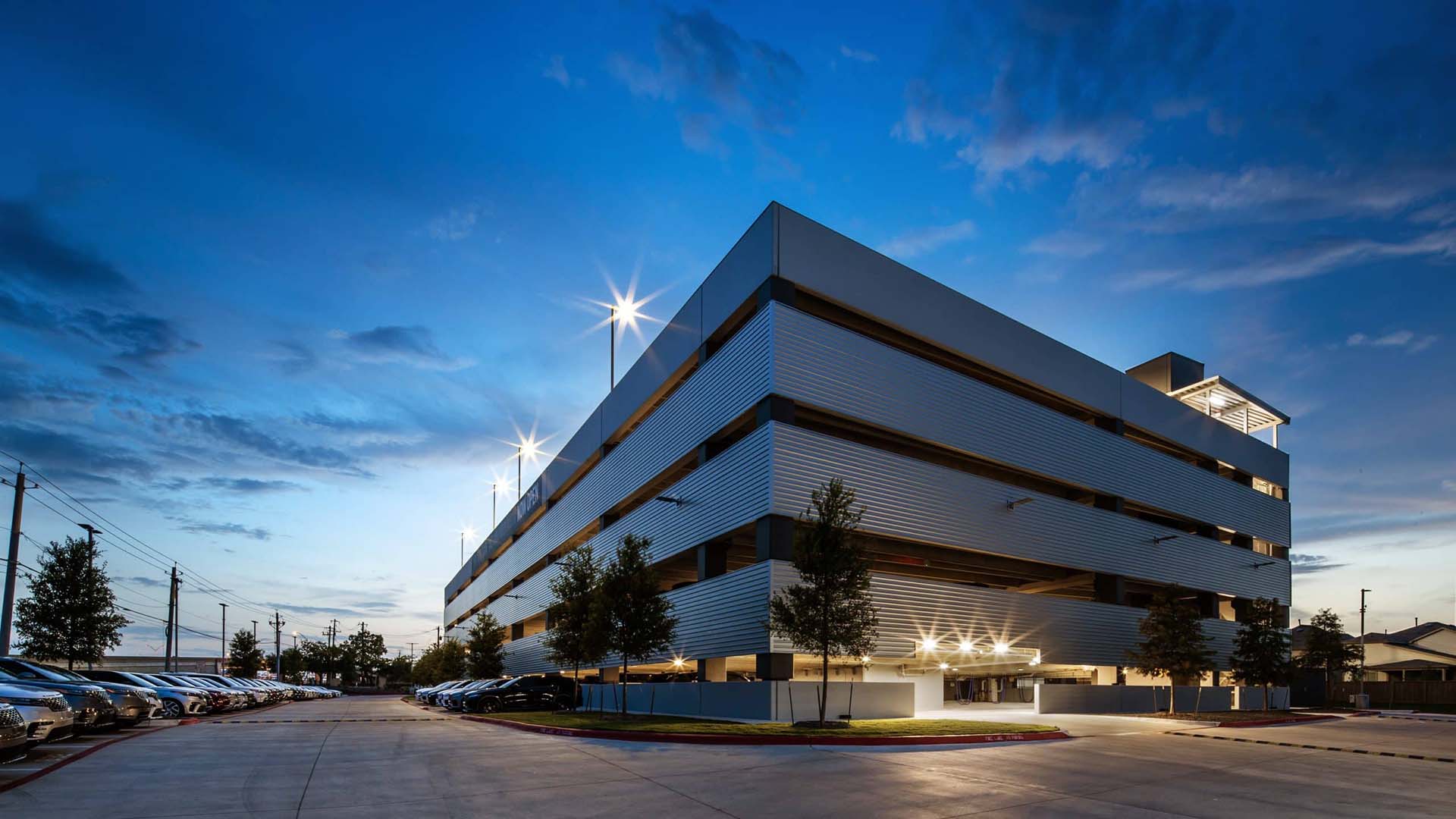 A multi-story parking garage is illuminated at dusk, with cars parked along its perimeter and streetlights visible in the background.