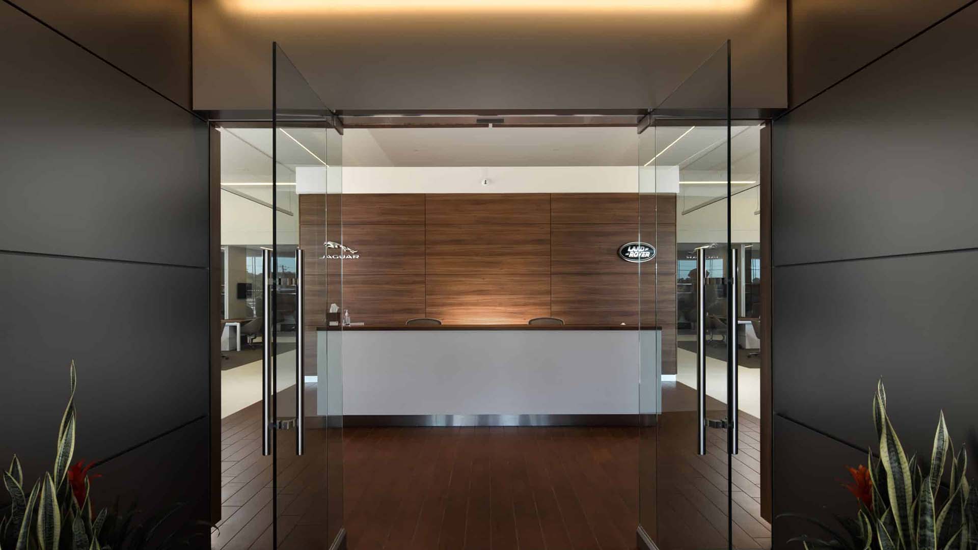 Modern office reception area with a white front desk, wooden wall panels, glass doors, and plants on either side of the entrance.
