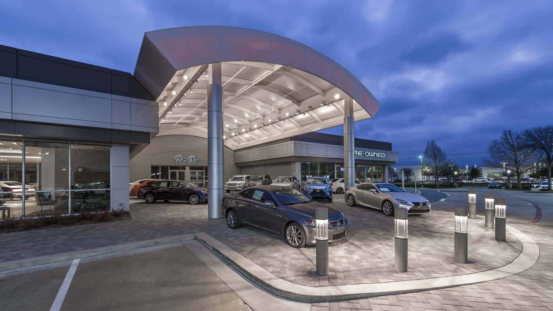A car dealership at dusk with several parked vehicles under a modern, well-lit canopy and a "Pre-Owned" sign visible in the background.