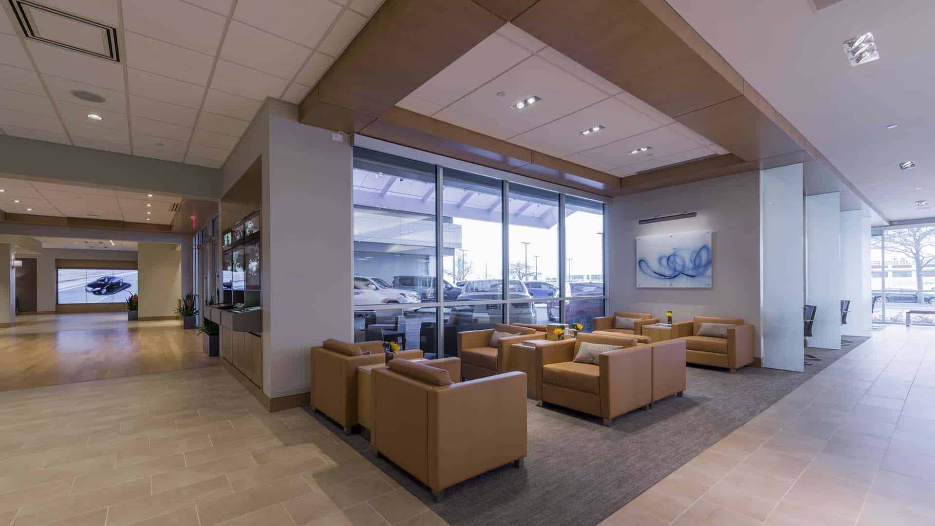 Modern waiting area with tan armchairs, small tables, and large windows overlooking parked cars; bright, clean, and spacious interior.