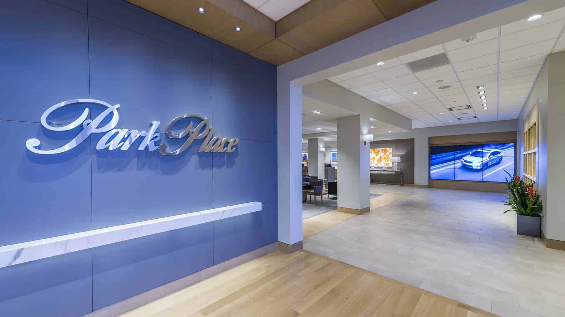 Modern dealership interior with "Park Place" sign on a blue wall, light wood and tile flooring, and a large display screen showing a car in the background.