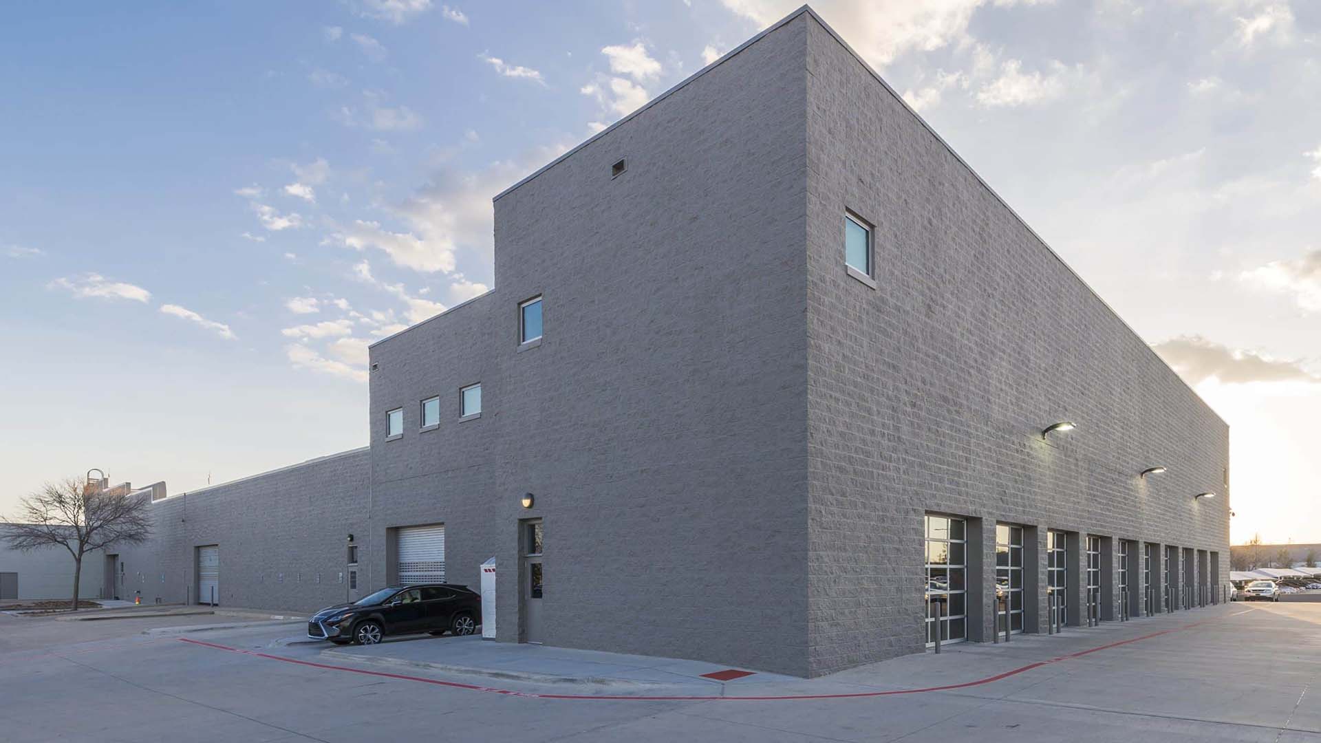 A large, modern industrial building with gray concrete walls, several garage doors, small windows, and a black car parked outside on a paved lot.