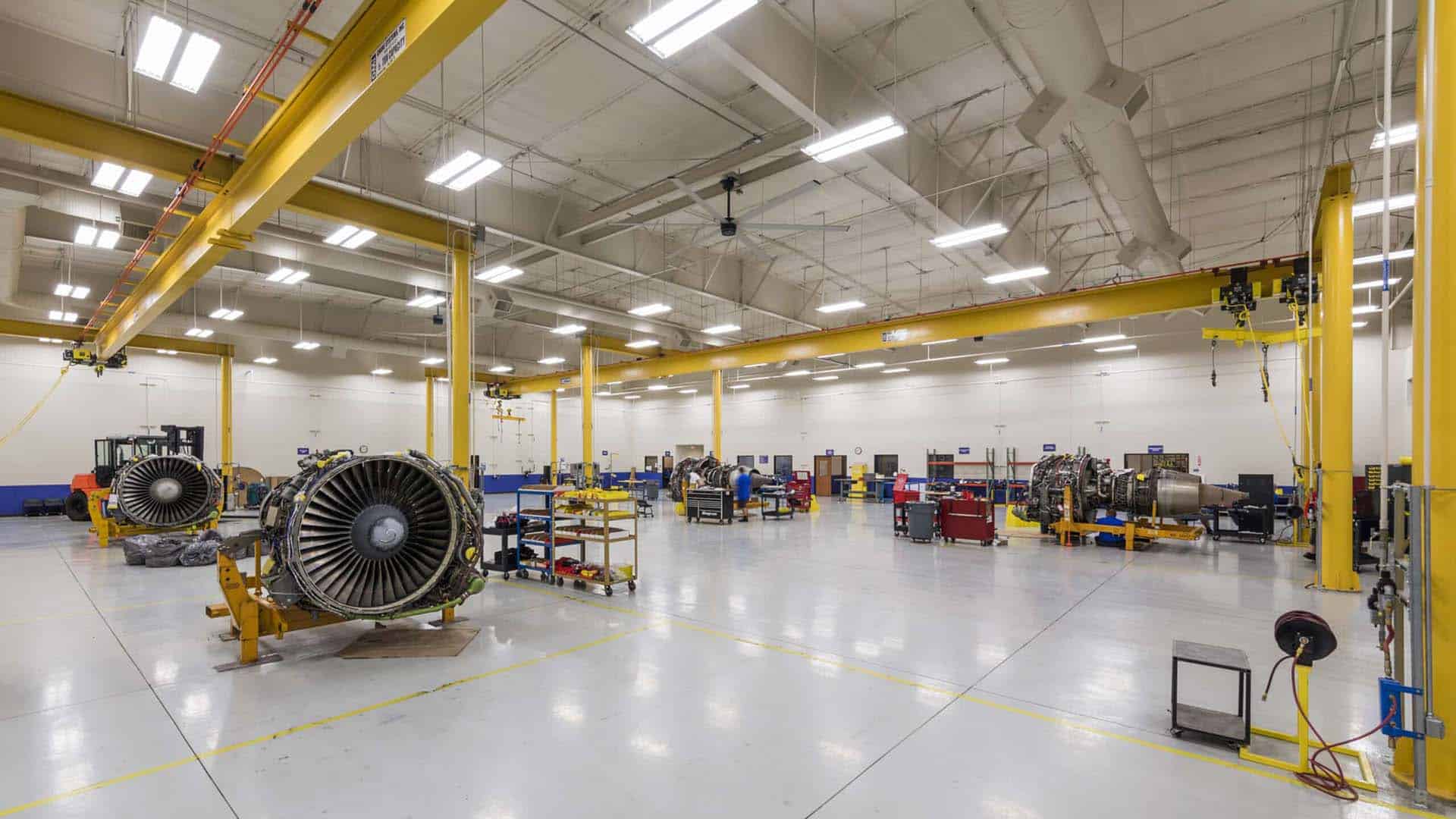 A spacious industrial workshop with large jet engines, yellow support beams, workstations, equipment carts, and bright overhead lighting.