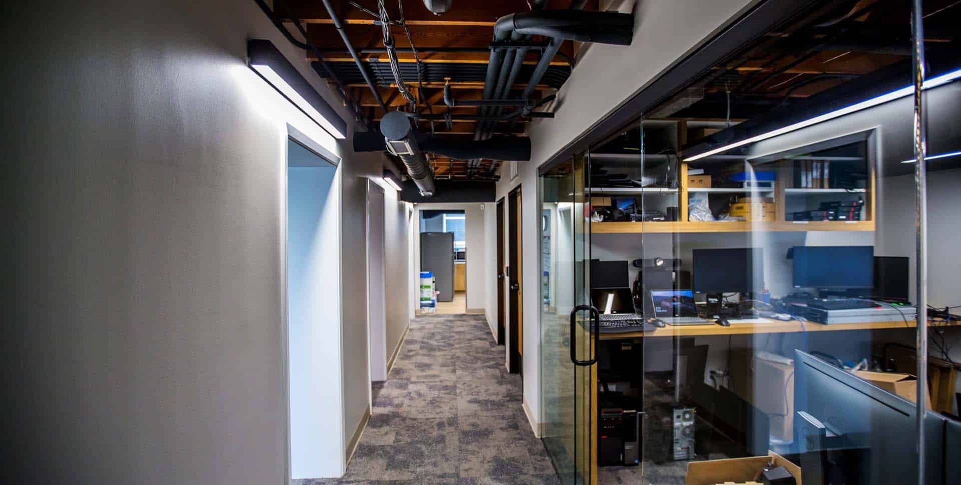 A modern office hallway with exposed ceiling pipes, carpeted floor, and glass-walled rooms containing desks and computer equipment.