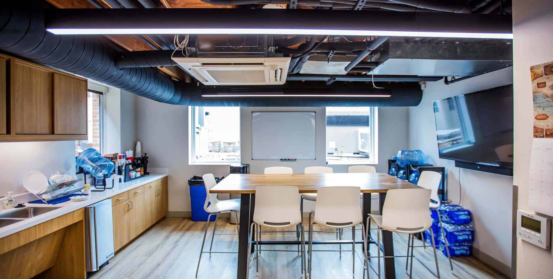 Modern office break room with a high table, white chairs, kitchen area, large screen, whiteboard, windows, and exposed ductwork on the ceiling.