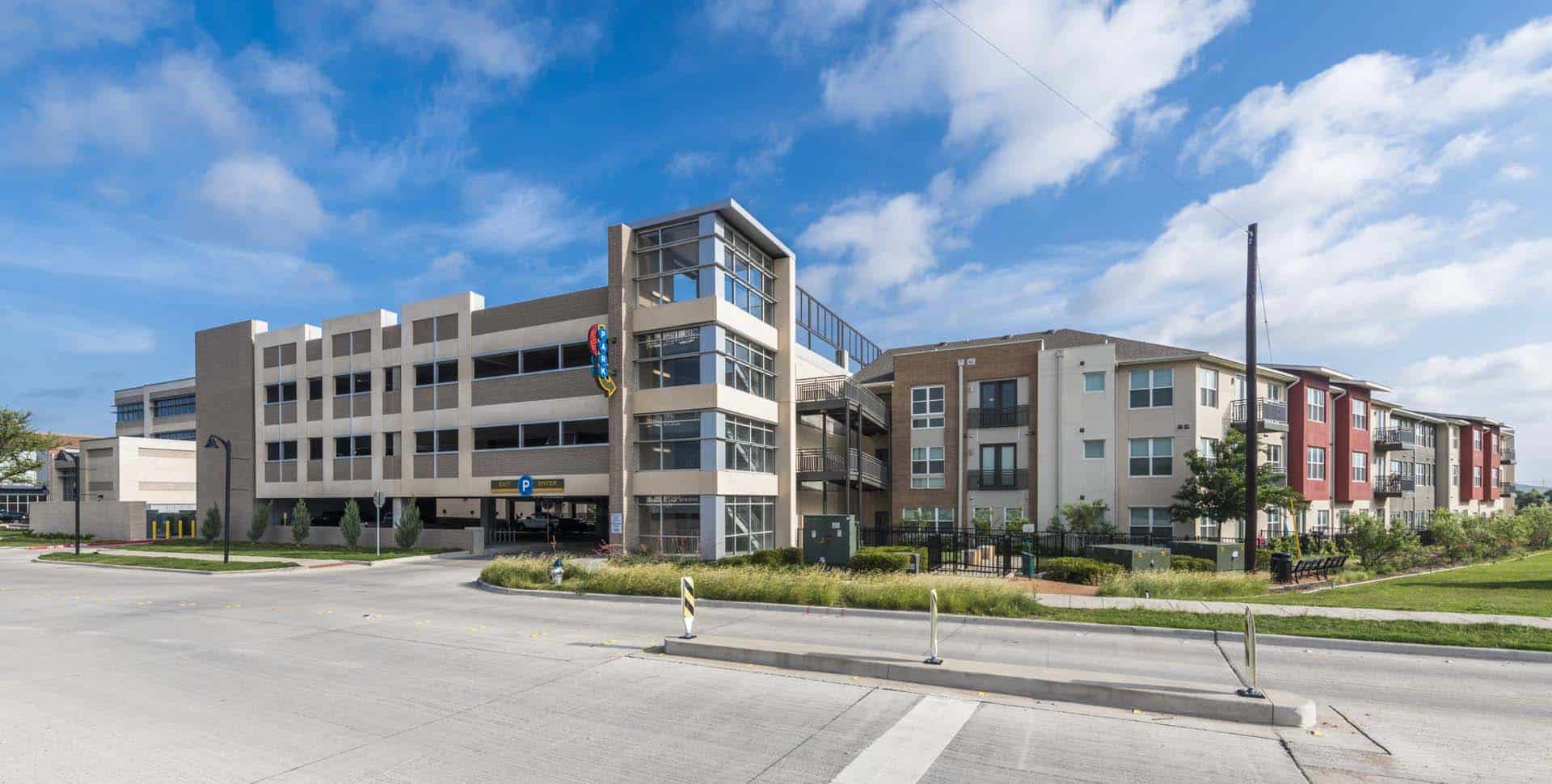 A multi-story parking garage is next to a modern apartment building on a sunny day with a partly cloudy sky.