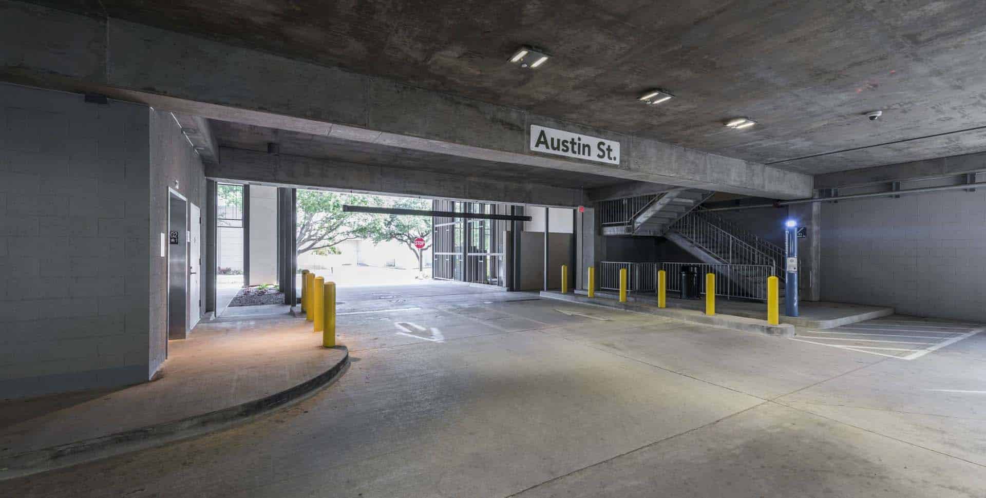 A concrete parking garage entrance with yellow bollards, an overhead sign reading "Austin St.," and a metal staircase on the right side.