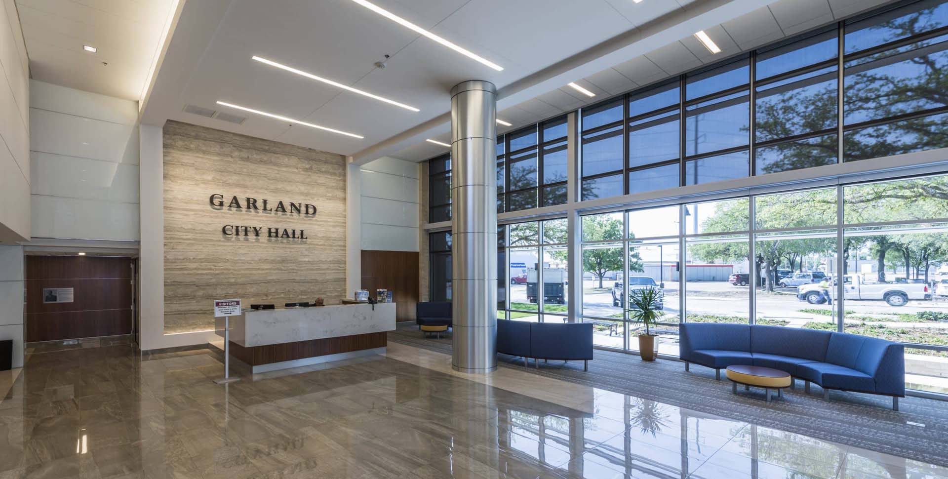 Modern lobby of Garland City Hall with large windows, tiled floor, reception desk, blue seating, and "Garland City Hall" sign on the wall.