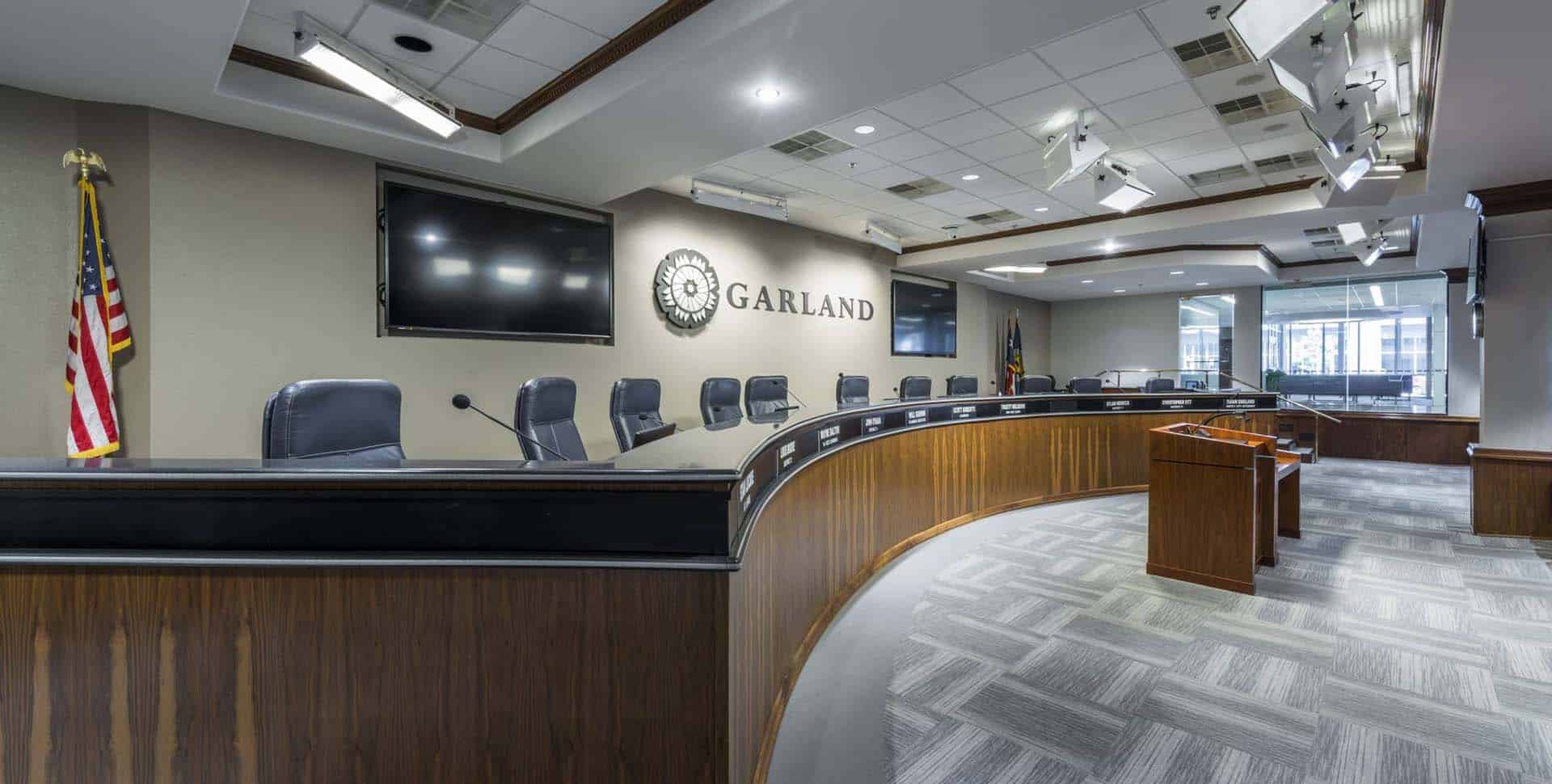 Empty city council chamber in Garland, Texas, featuring a curved wooden desk with microphones, chairs, USA and Texas flags, and multiple monitors on the walls.