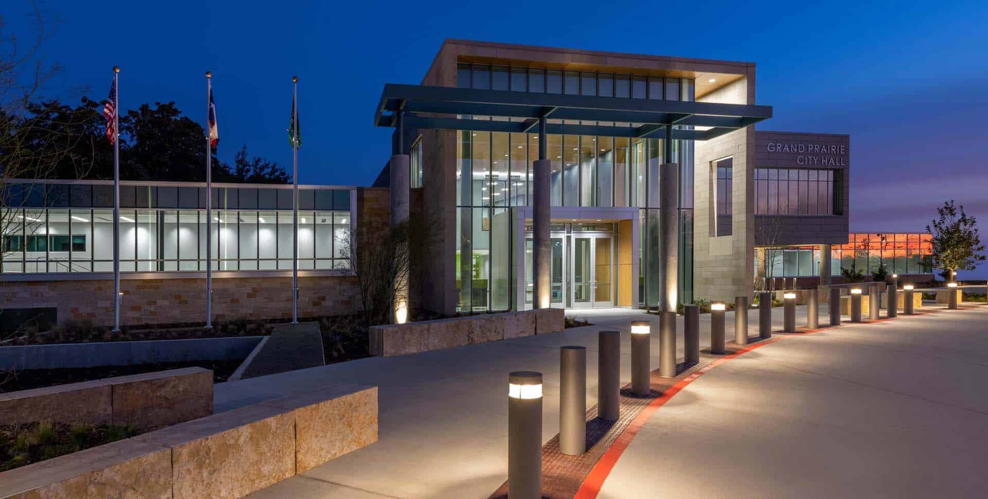 Modern building with large glass windows and illuminated entrance, labeled "Grand Prairie City Hall," surrounded by lighted bollards and flagpoles at dusk.