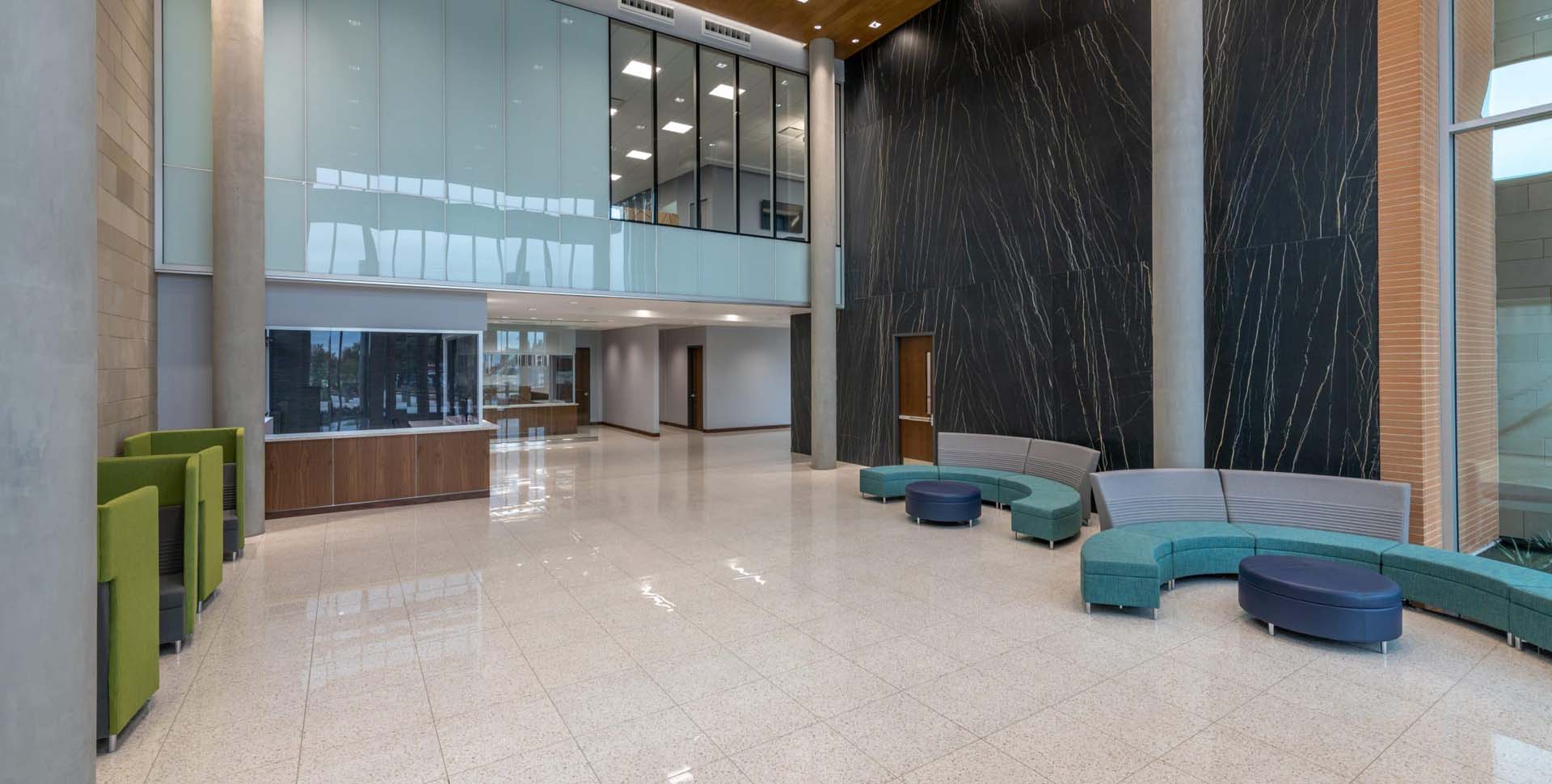 Spacious modern lobby with tiled floor, glass balcony, black accent wall, curved seating, green booths, and natural light from large windows.