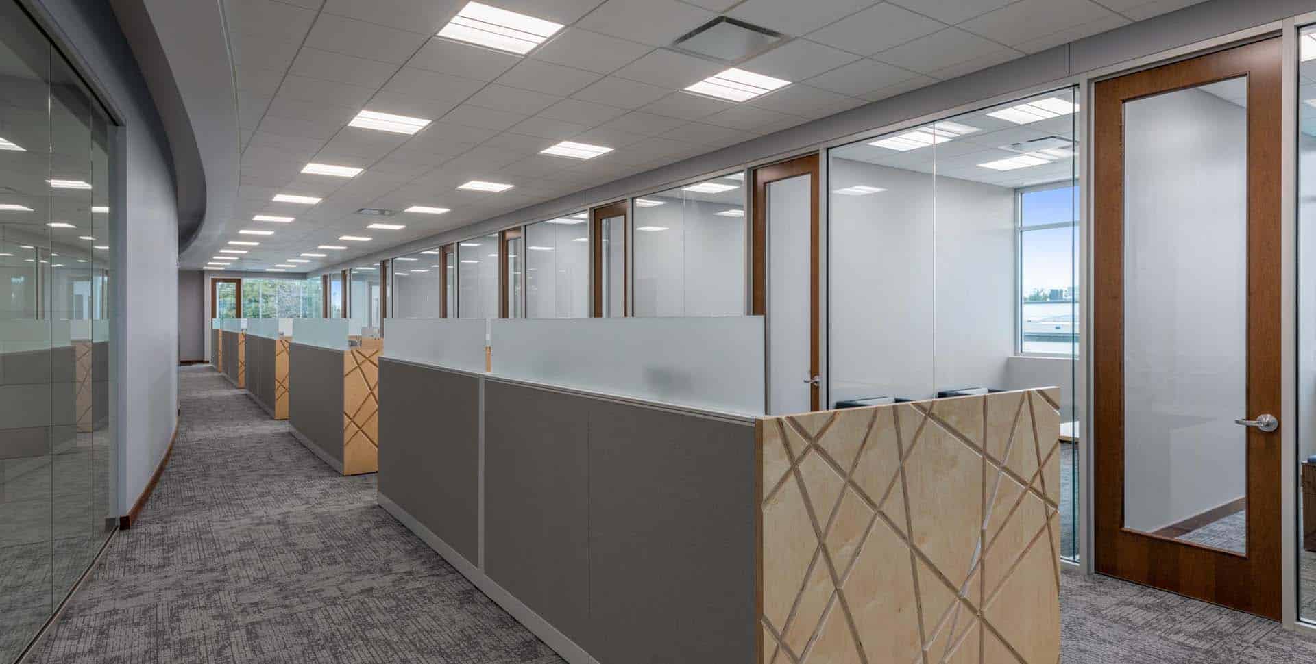 Modern office space with carpeted floors, glass-walled private offices on the right, and cubicle workstations lining the corridor on the left.