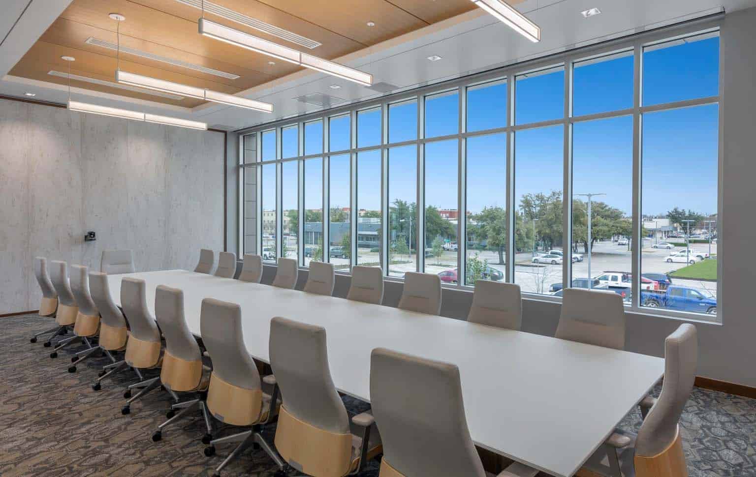 A modern conference room with a long white table, many beige chairs, and large windows overlooking a parking lot and trees.