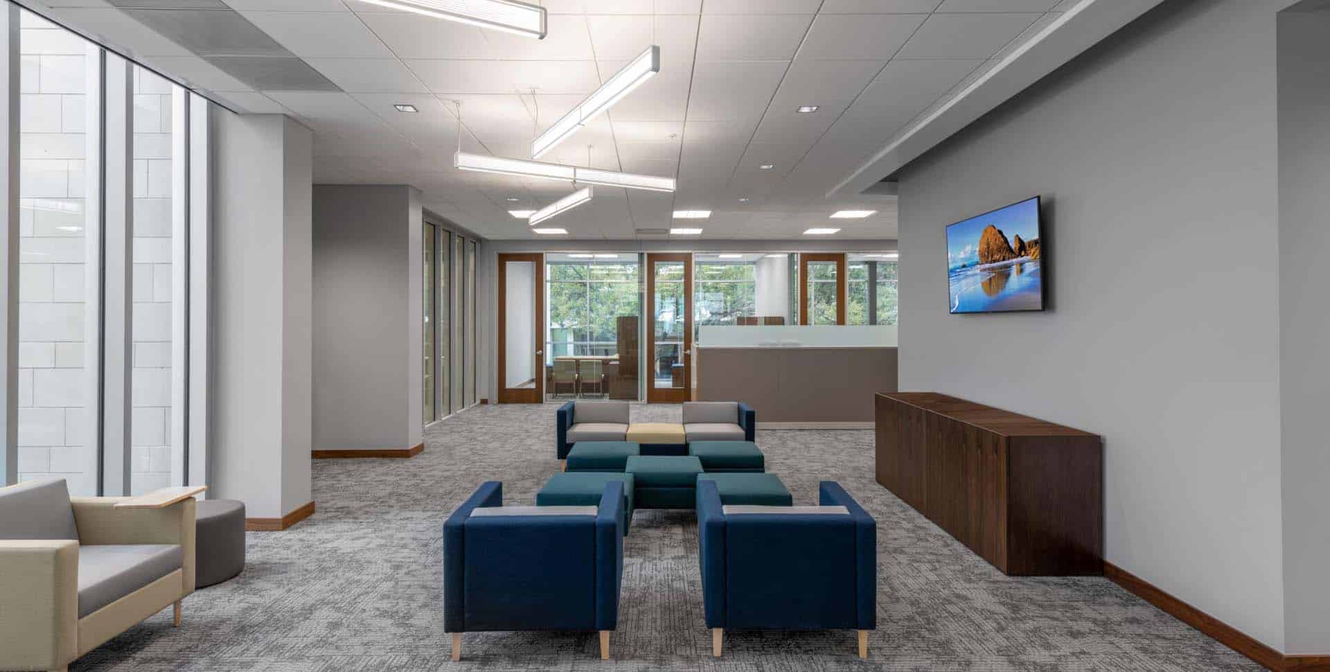 Modern office waiting area with blue and beige chairs, a wooden console, wall-mounted TV, and large windows letting in natural light.