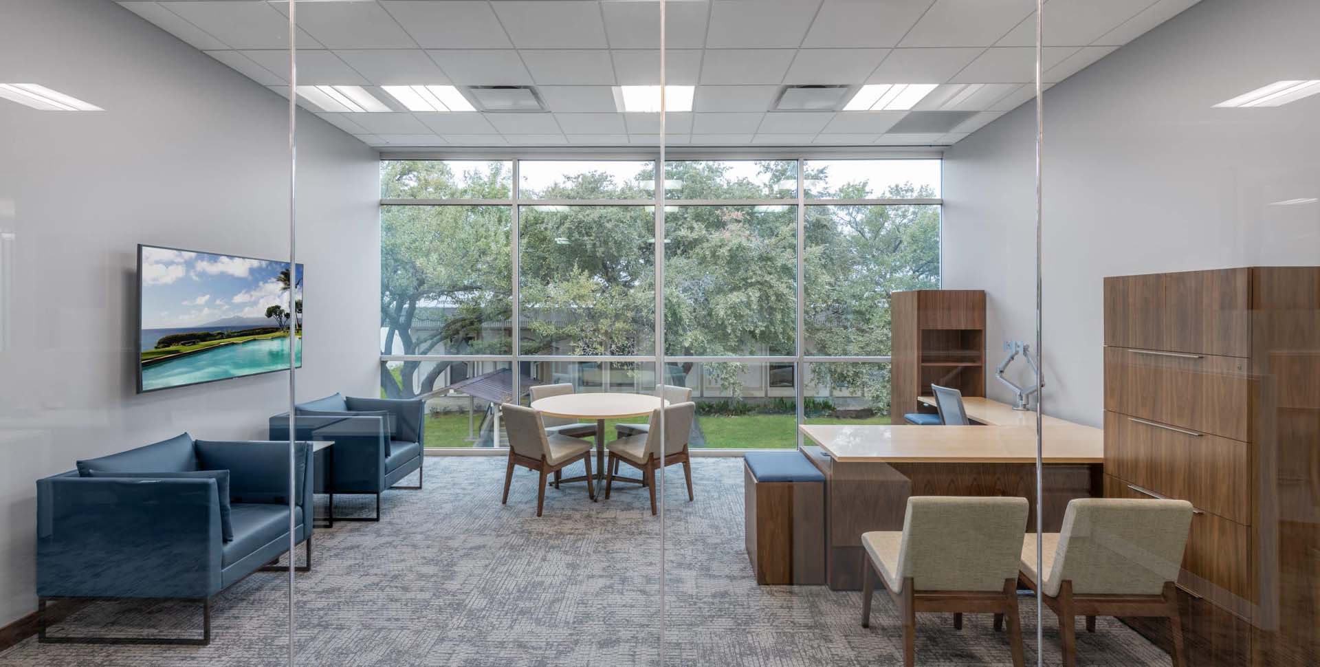 Modern office with a large window, wooden desk, filing cabinets, round meeting table with chairs, two blue armchairs, TV on the wall, and trees visible outside.