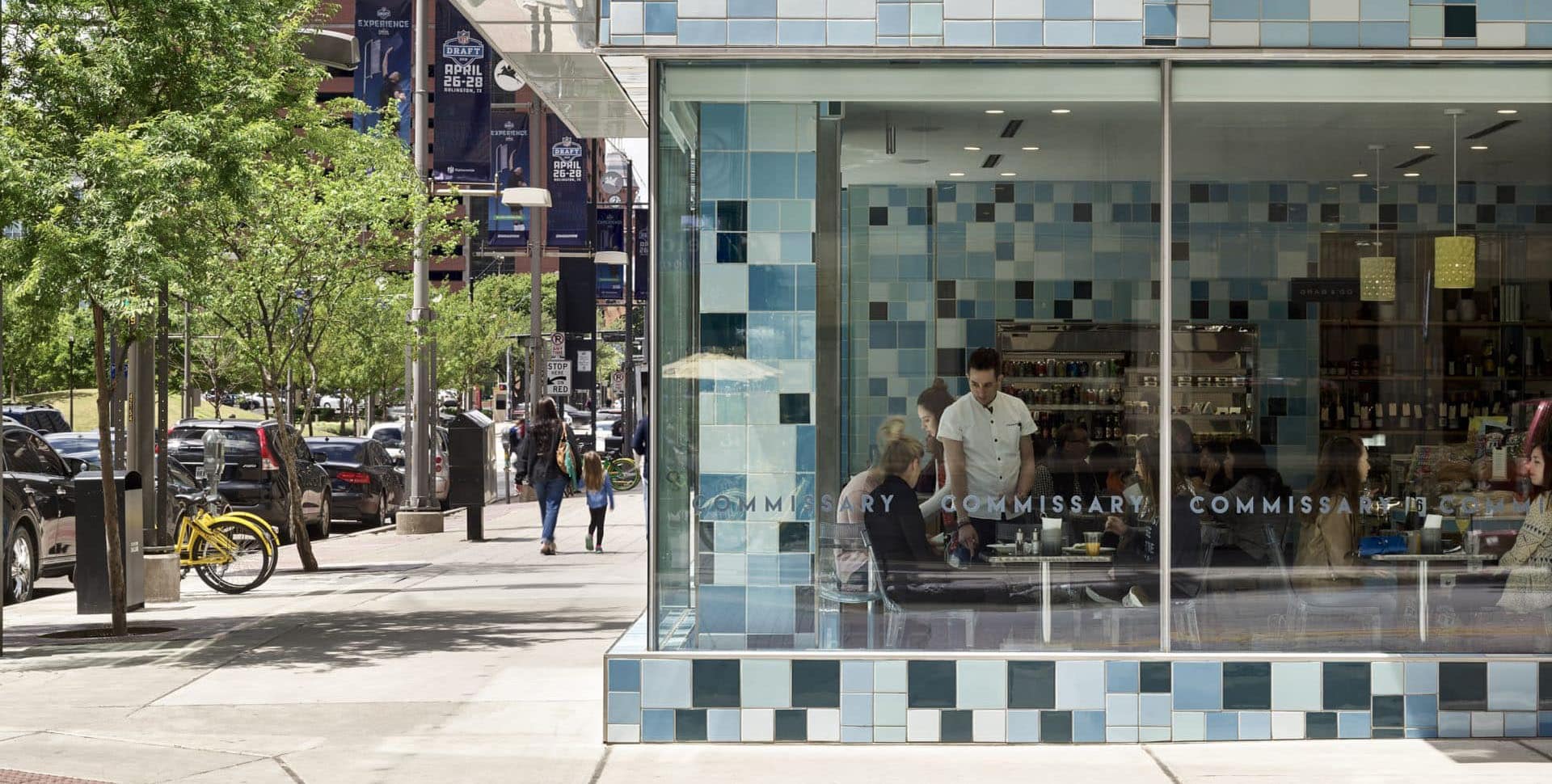 People are seated inside a modern café with large windows and blue tiles, while pedestrians and parked cars line the tree-lined street outside.