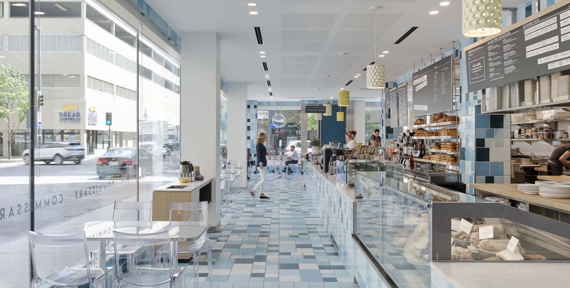 Modern bakery and café interior with blue tile flooring, glass display cases of baked goods, menu boards, and large windows facing a city street.