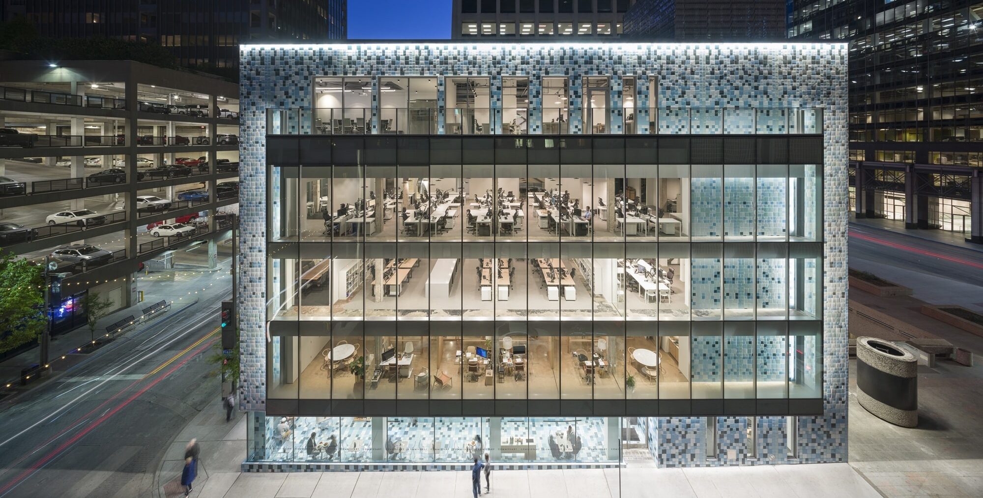 A modern office building with a façade of blue and gray square tiles and large glass windows revealing brightly lit workspaces inside. Several floors of desks and meeting areas are visible. Surrounding the building are tall skyscrapers, a multi‑level parking garage filled with cars, and city streets illuminated at night.