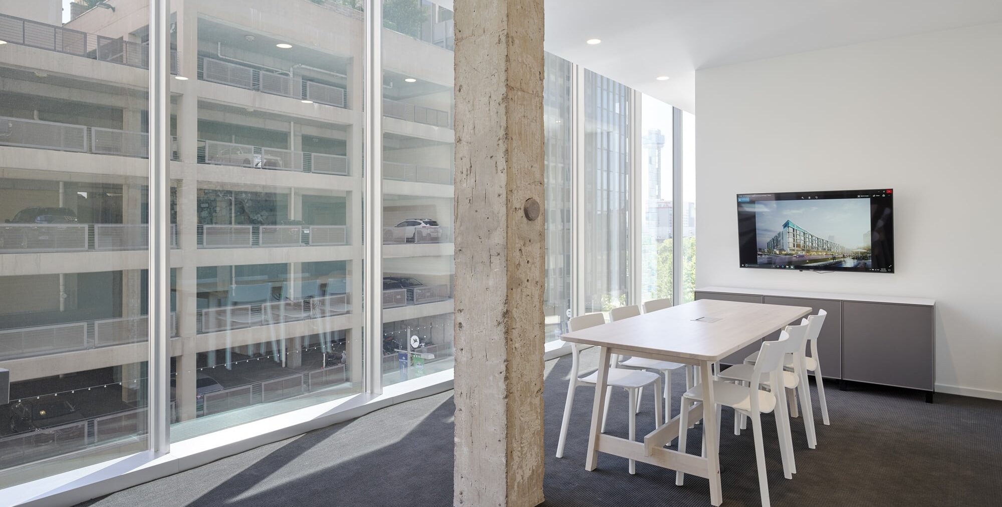 A minimalist meeting room with a light wooden table surrounded by white chairs on dark carpet. A rough concrete column stands near the center of the space. Floor‑to‑ceiling windows show a parking structure and tall buildings outside. A wall‑mounted monitor displays an architectural image.