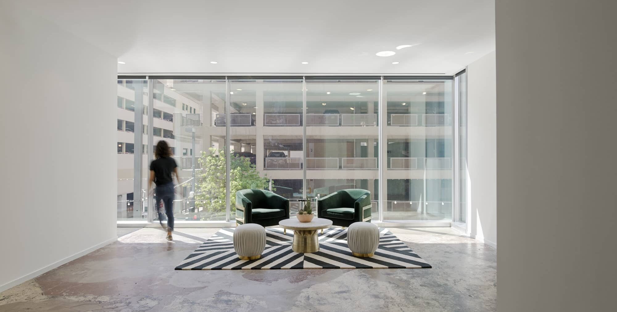 A modern seating area with two dark green armchairs, a round white coffee table with a gold base, and two pleated white ottomans arranged on a black‑and‑white striped rug. Floor‑to‑ceiling windows overlook a parking structure and greenery. A person walks toward the window, slightly blurred in motion.
