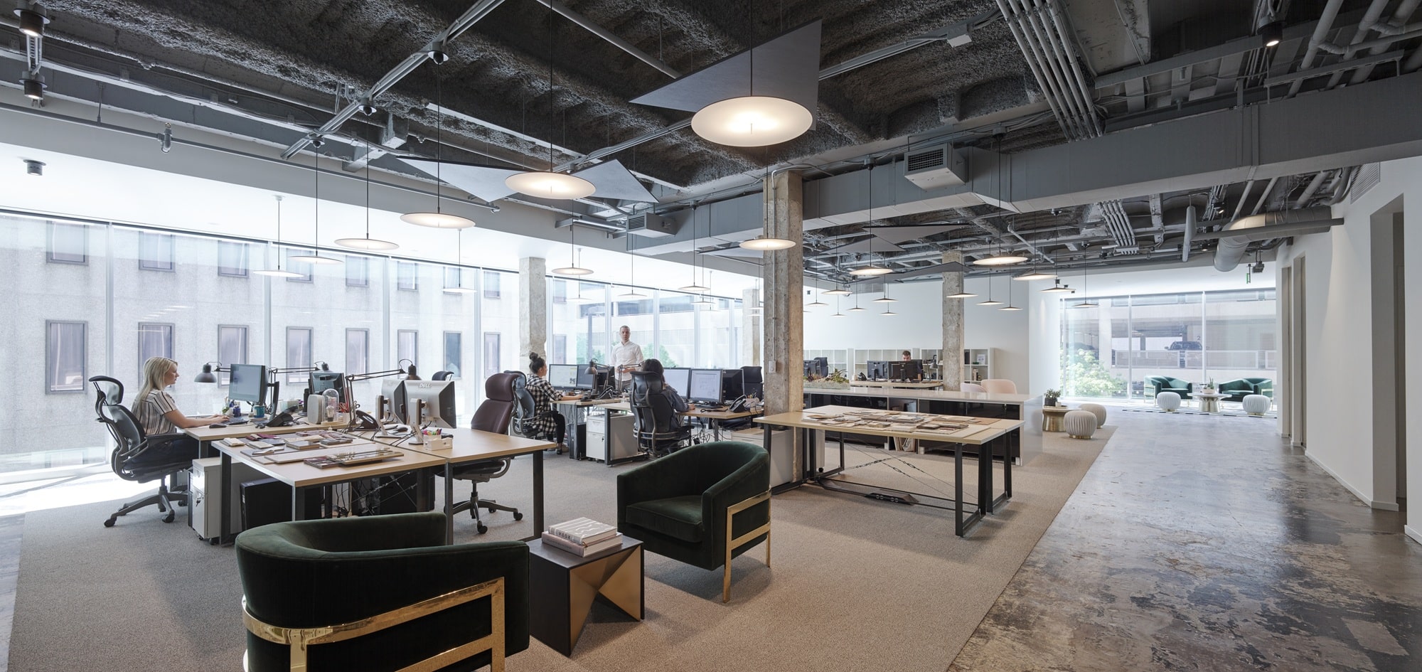 A contemporary open office with desks, computers, and people working. Large windows bring in natural light. The ceiling features exposed ducts and pipes with modern lighting. Lounge seating with green upholstery and gold accents sits near polished concrete and carpeted flooring.