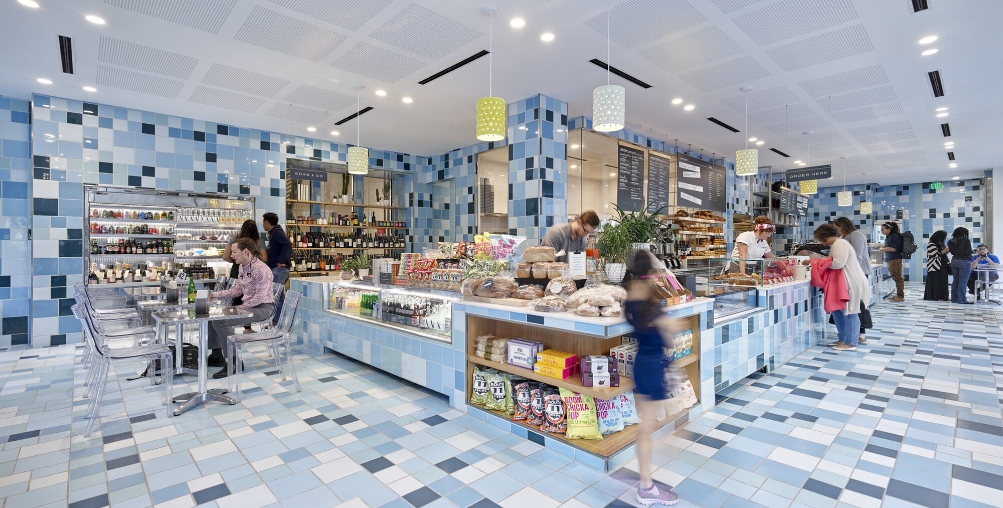 A bright café with blue‑and‑white mosaic tile walls and floors. A central counter displays baked goods and packaged snacks, with shelves of wine bottles behind it. Green and white pendant lights hang overhead. People sit at transparent tables or stand in line to order.