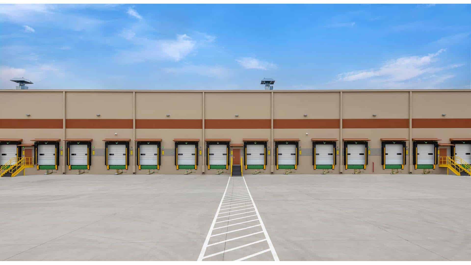 A row of closed loading dock doors with yellow stairs on a large industrial warehouse, set against a blue sky with light clouds.