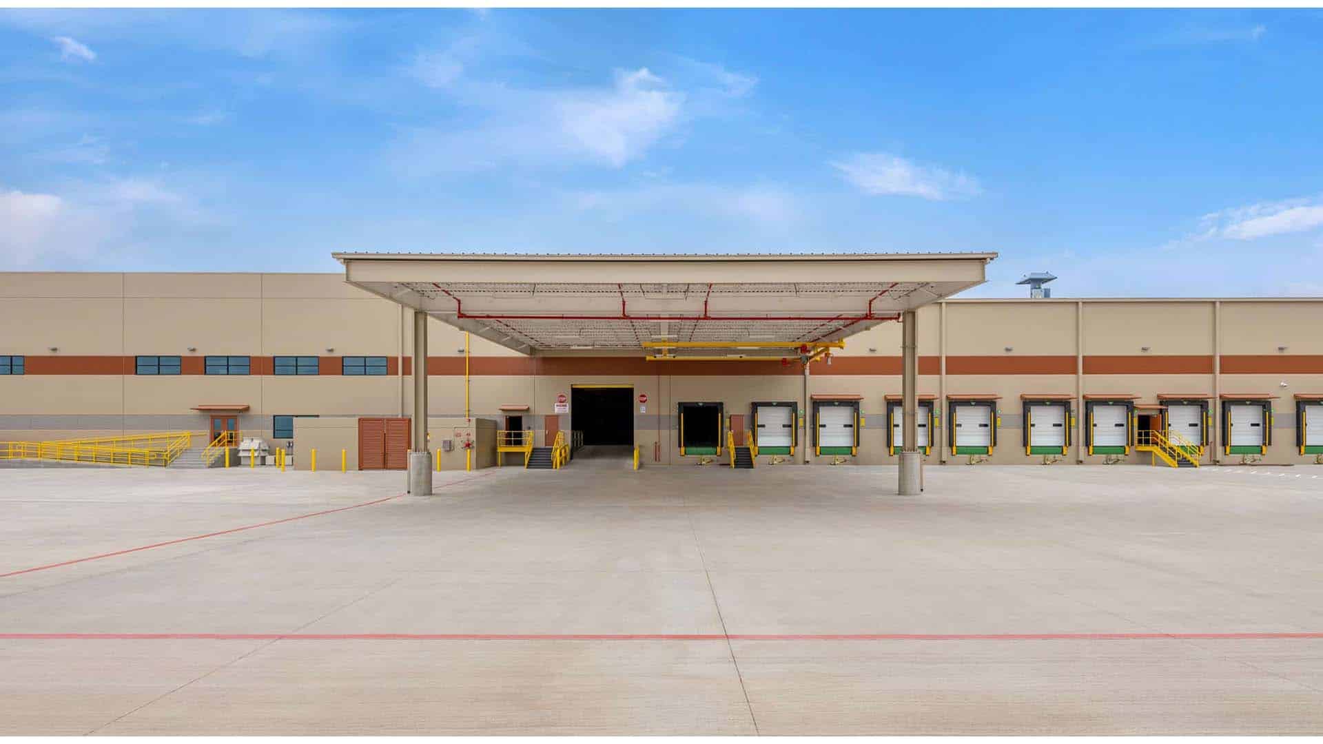 A large industrial warehouse loading dock with multiple bay doors, covered canopy, yellow railings, and a wide concrete driveway under a blue sky.