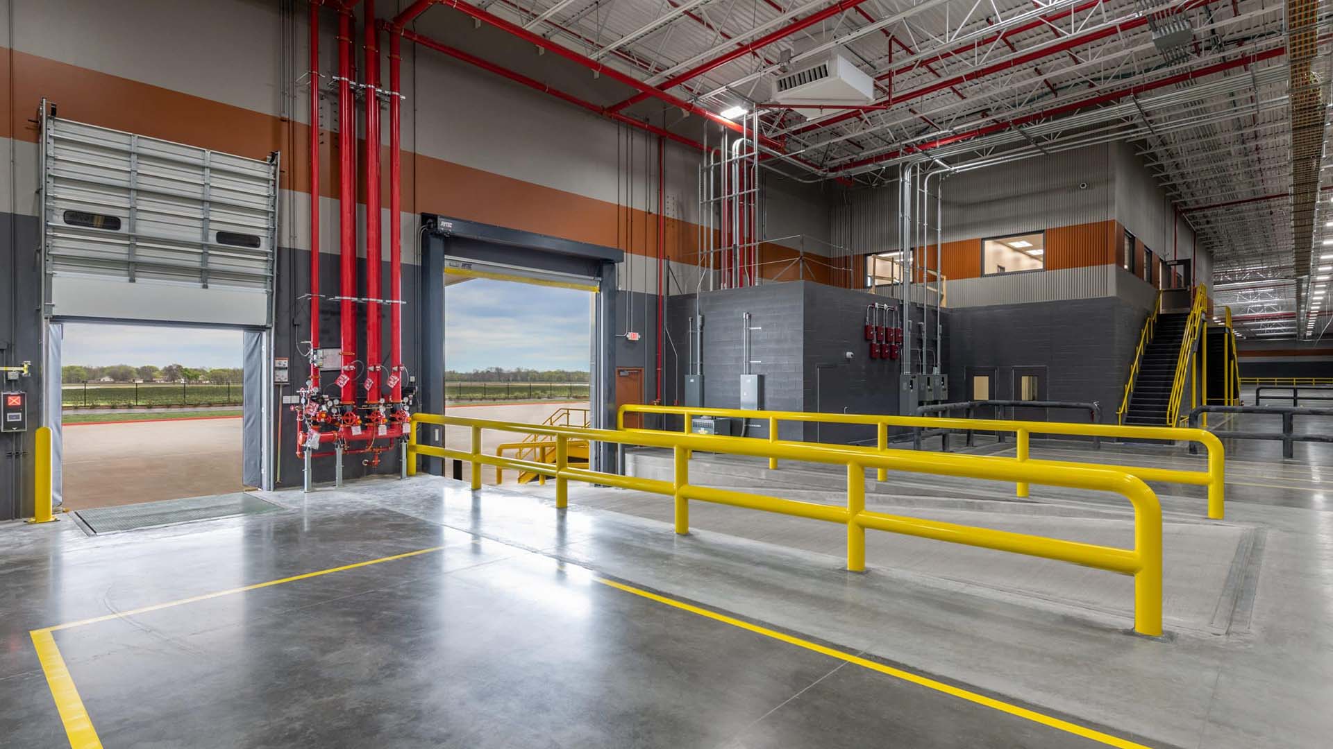 Interior of a modern warehouse with yellow safety railings, loading dock doors open to outside, concrete floors, and visible fire safety systems.