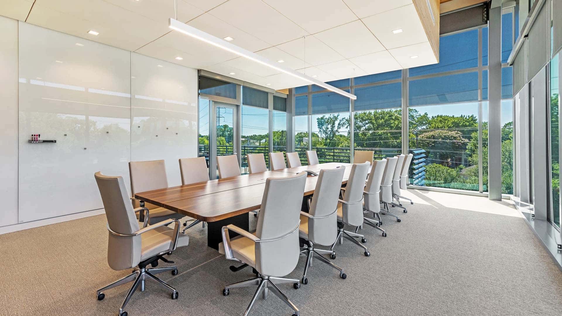 Modern conference room with a long wooden table, twelve beige chairs, large windows, and a whiteboard wall; greenery visible outside.