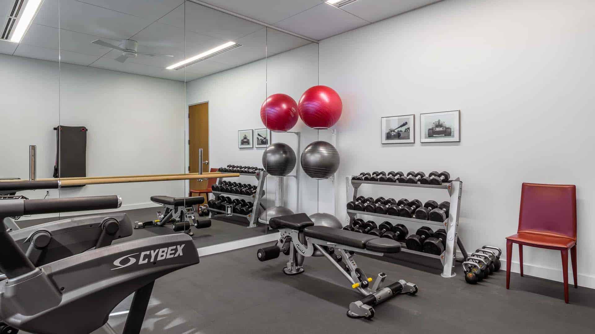 A small gym room with free weights, benches, exercise balls, a Cybex machine, a mirror wall, and framed photos; a red chair sits in the corner.