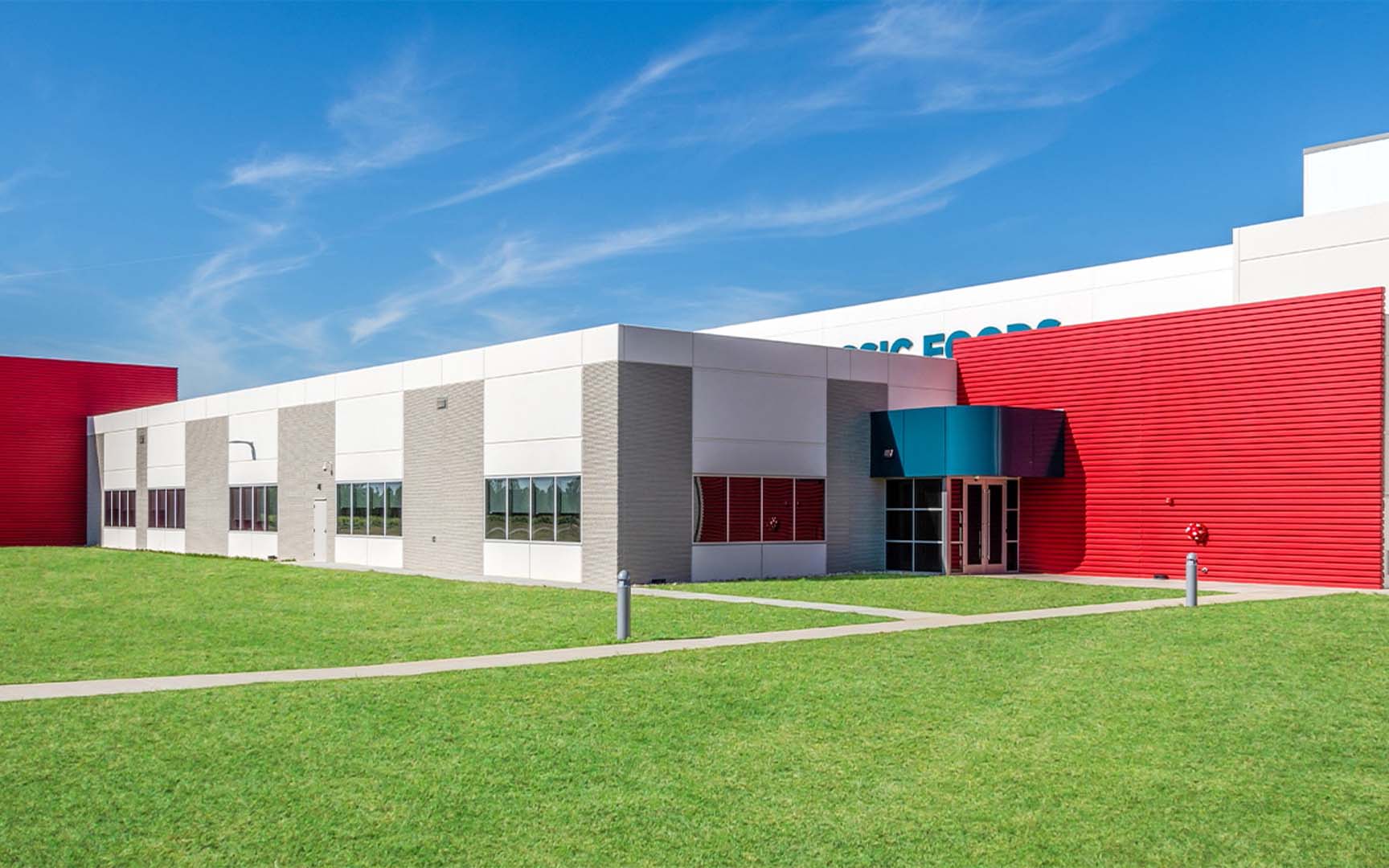 Modern industrial or commercial building with white, gray, and red exterior panels, large windows, blue entrance canopy, and green lawn under a blue sky.