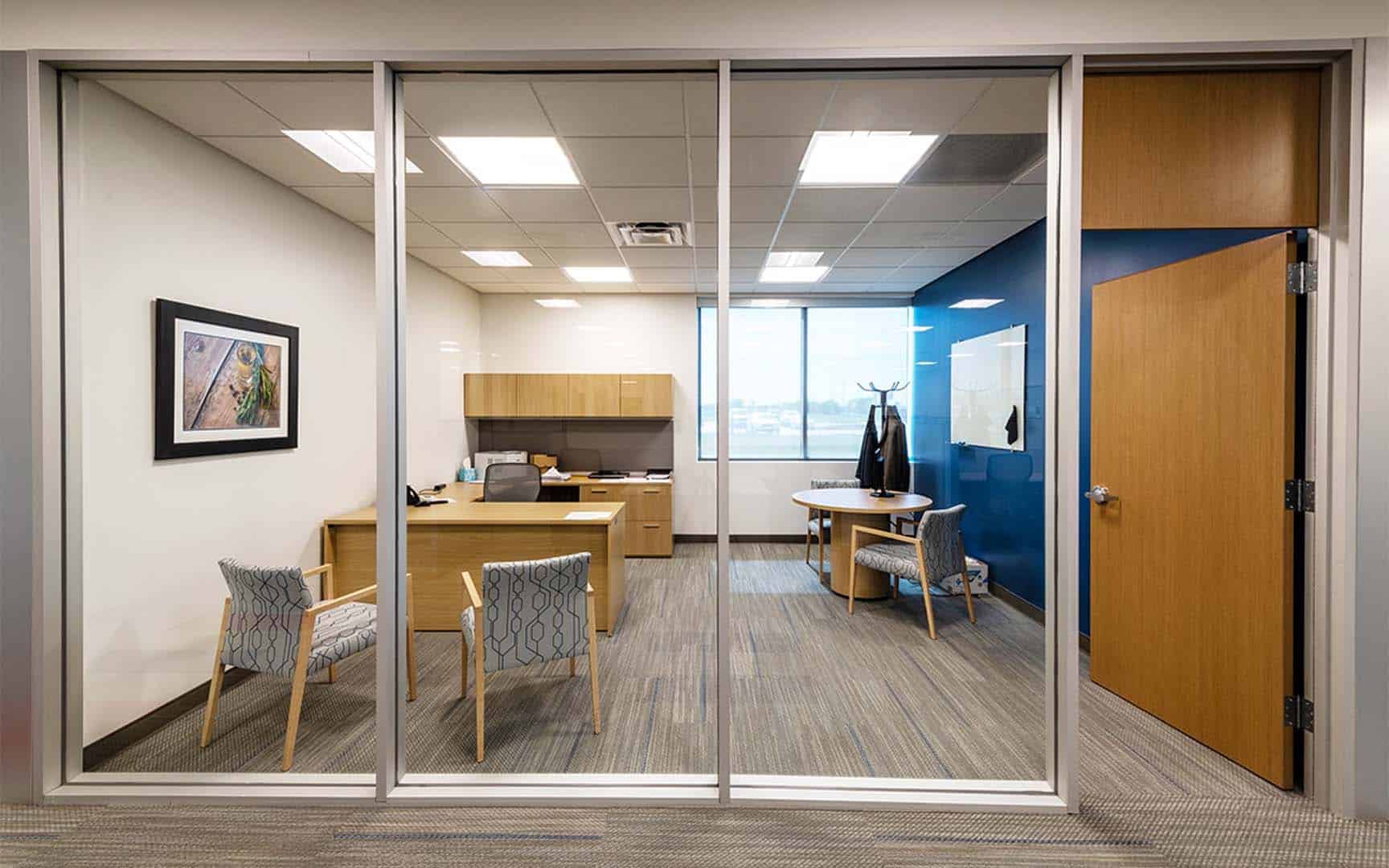 Modern office with a desk, two guest chairs, cabinets, a window, a coat rack, and a framed picture on the wall, viewed through glass walls and a door.