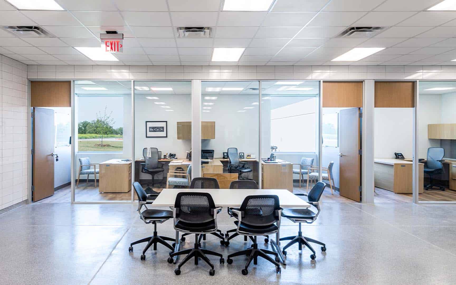 A modern office space with glass-walled private offices and an open area featuring a table with six chairs on wheels.