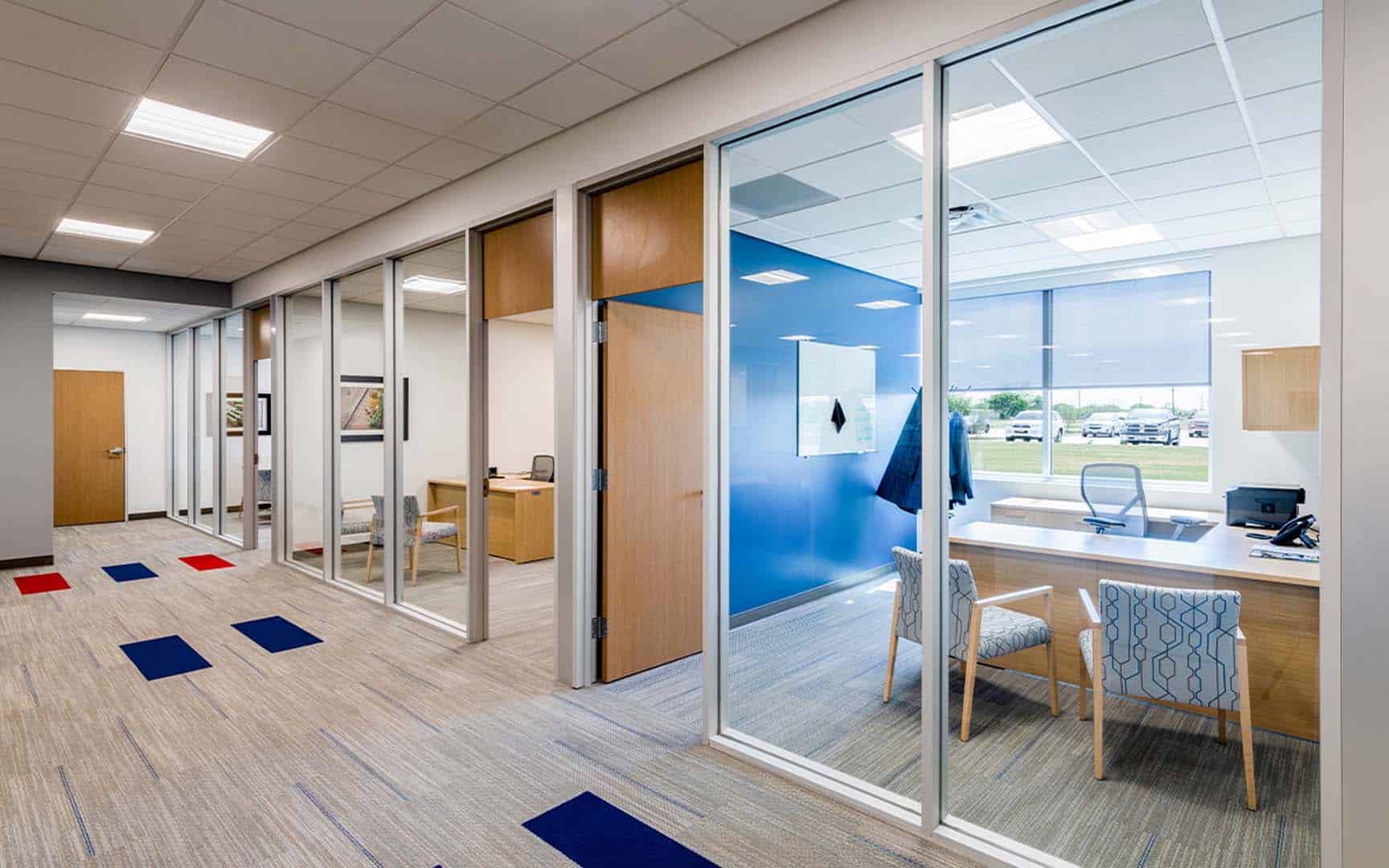 Modern office hallway with glass-walled private offices, carpeted floor with colored square accents, and desks and chairs visible inside the offices.