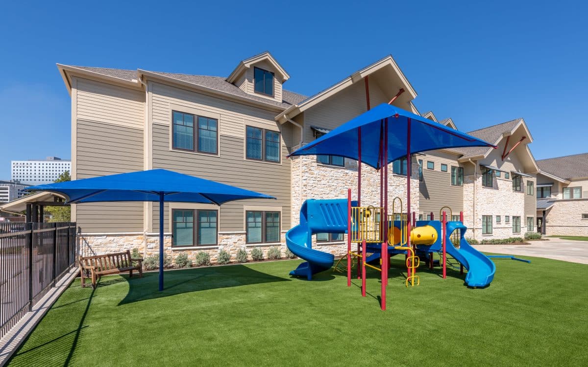 A playground with a blue slide and umbrella in front of an apartment building.
