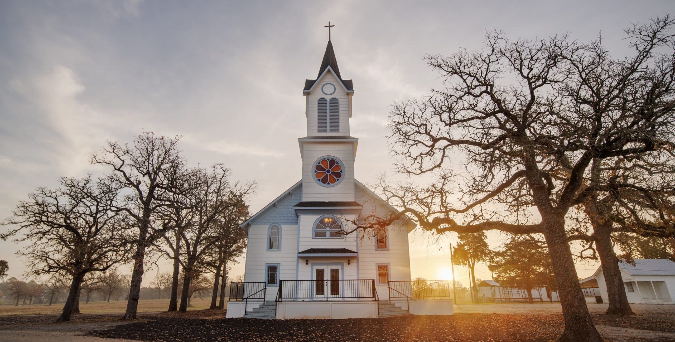 A white wooden church with a tall steeple, double front doors, arched windows, and a circular stained‑glass window. Leafless trees surround the building, and warm low sunlight casts long shadows across the ground.