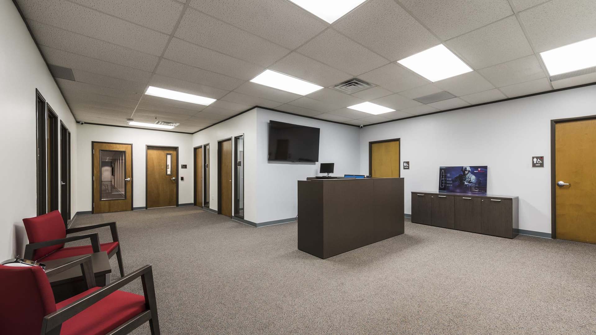 A modern office waiting area with red chairs, a dark reception desk, wall-mounted TV, and several wooden doors along white walls.